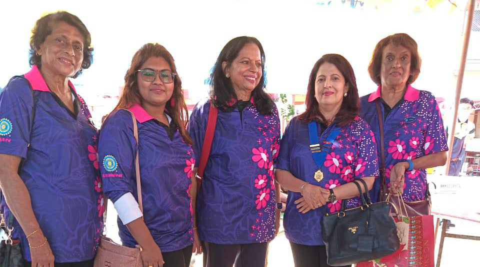 Five adults wearing matching purple shirts with pink floral designs stand together outdoors on a sunny day, some holding bags and smiling.
Description generated by AI