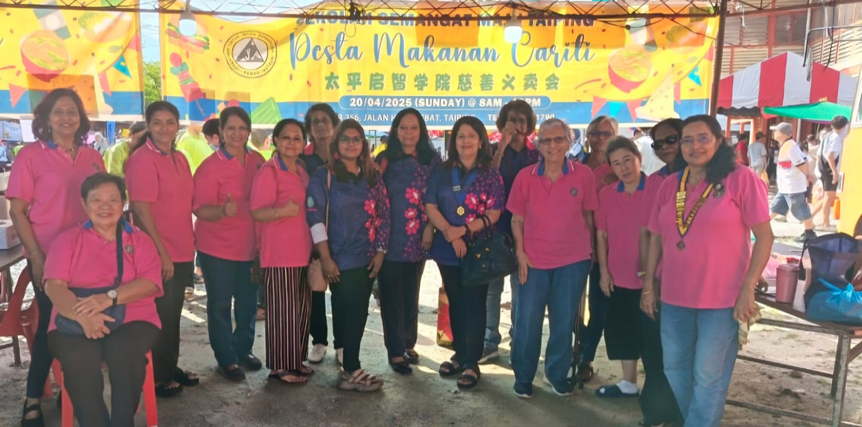 A group of people wearing pink and blue shirts stand together under a tent with a colorful banner promoting a cultural food festival in the background.
Description generated by AI