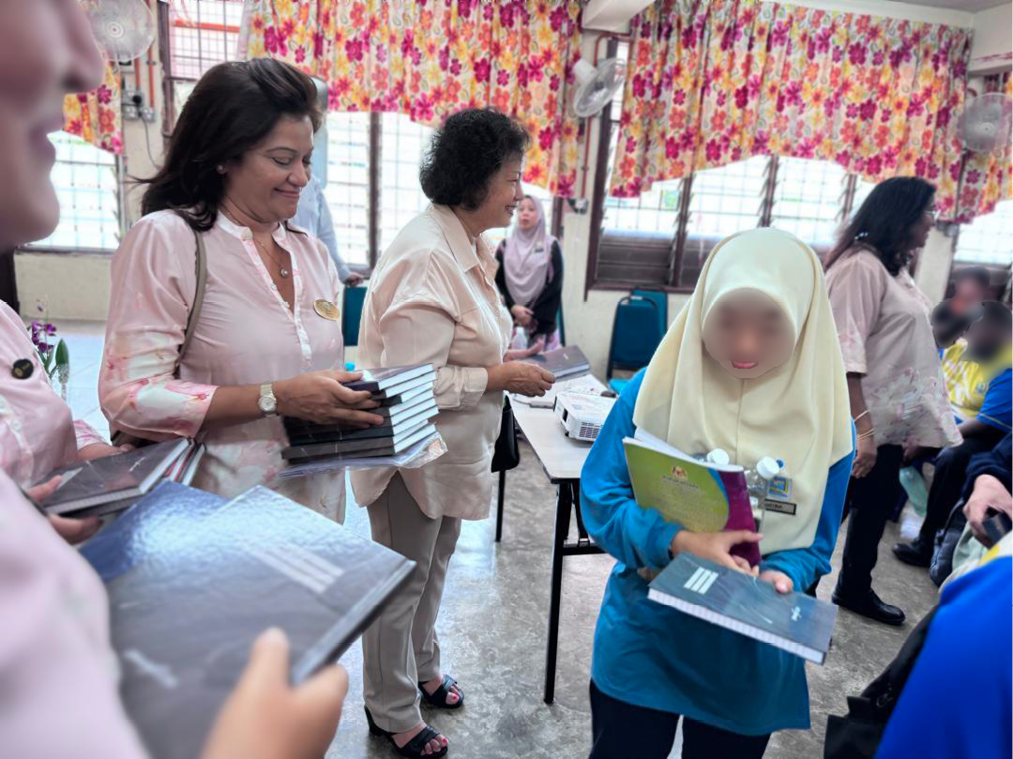 People in a room are distributing and receiving books, with some wearing light-colored tops and one person in a blue outfit with a light-colored headscarf holding books.
Description generated by AI