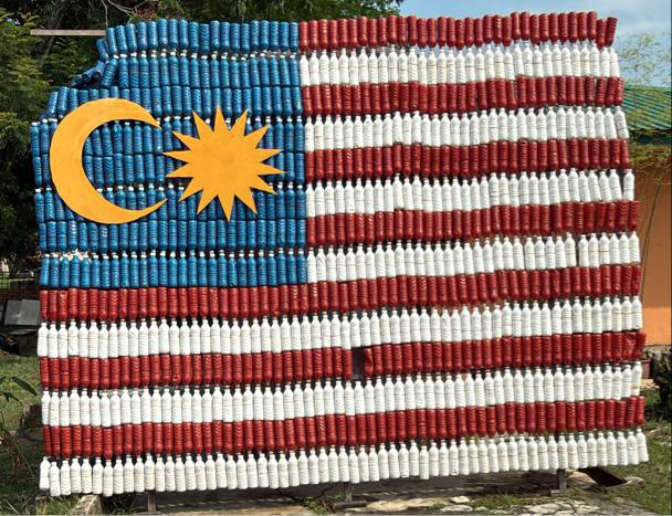 A large Malaysian flag made from stacked plastic bottles is displayed outdoors against a backdrop of trees and sky.
Description generated by AI