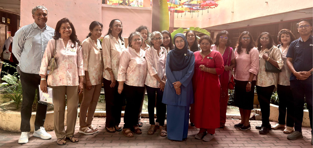 A group of people standing together in a courtyard with colorful umbrellas hanging above, some wearing matching light-colored tops and one person in a blue outfit standing in the center.
Description generated by AI