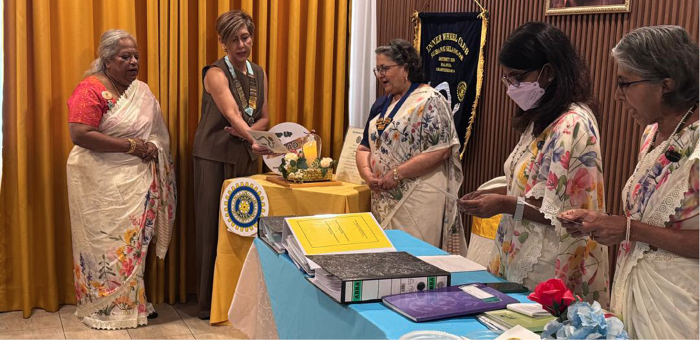 Five adults wearing traditional clothing stand around tables with documents and flowers in a formal setting with yellow curtains and a banner in the background.

Description generated by AI