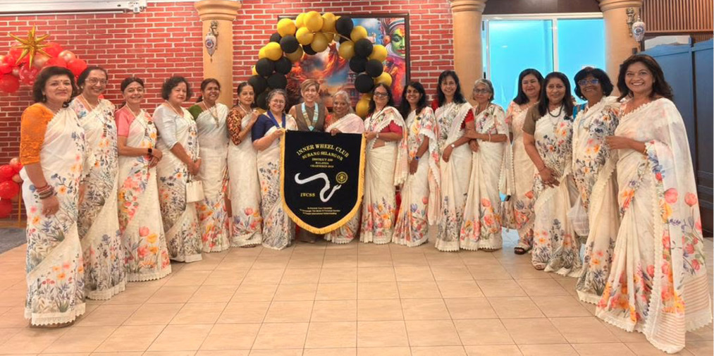 A group of people dressed in white sarees with floral patterns stand indoors in a semi-circle around a banner with black and yellow balloons above it.

Description generated by AI