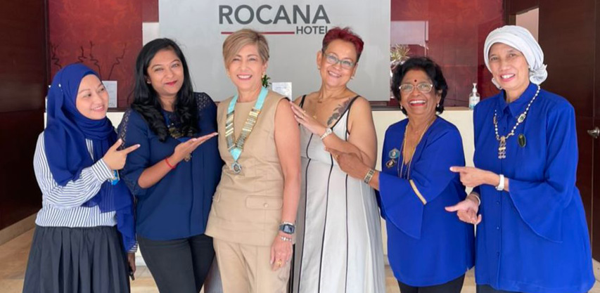 Six adults stand smiling in front of a Rocana Hotel sign with five of them pointing towards the person in the center who wears a beige outfit and a ceremonial chain.
Description generated by AI