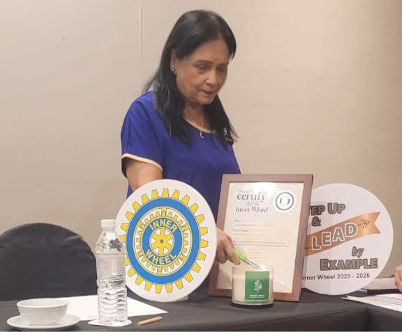 A person in a blue shirt is standing behind a table with a water bottle, a cup and saucer, a pencil, and two circular signs, one with a Rotary International logo and the other with the text "Step Up & Lead by Example," while holding a framed certificate.

Description generated by AI