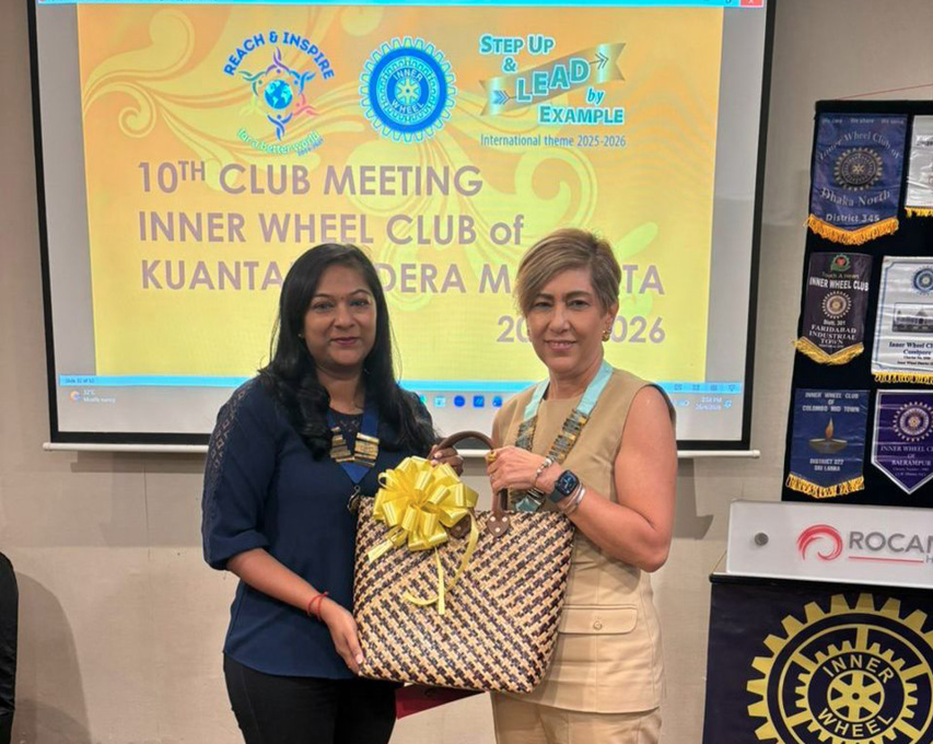 Two adults stand indoors in front of a presentation screen and banners, holding a large gift bag with a yellow bow, during a club meeting event.
Description generated by AI
