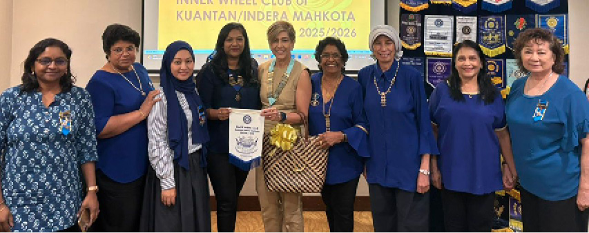 A group of adults standing indoors in front of a screen and banners, some holding a flag and a gift bag, dressed mostly in blue attire, at a club meeting event.
Description generated by AI