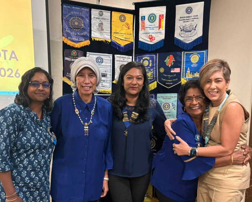 Five adults stand closely together smiling in front of a display of colorful banners, with one person hugging another while the others stand side by side wearing casual and semi-formal clothing.
Description generated by AI