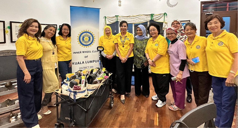 A group of people wearing yellow shirts and casual clothing stand indoors around a cart filled with various items next to a banner for the Inner Wheel Club of Kuala Lumpur.
Description generated by AI
