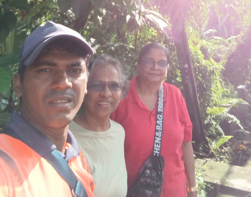 Three adults stand close together outdoors with greenery and sunlight in the background, one wearing a cap and an orange shirt, another in a light-colored shirt, and the third in a red shirt with a crossbody bag.
Description generated by AI