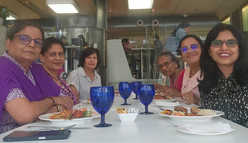 Six adults sitting around a table in a restaurant with plates of food and blue drinking glasses, smiling and looking toward the camera.
Description generated by AI