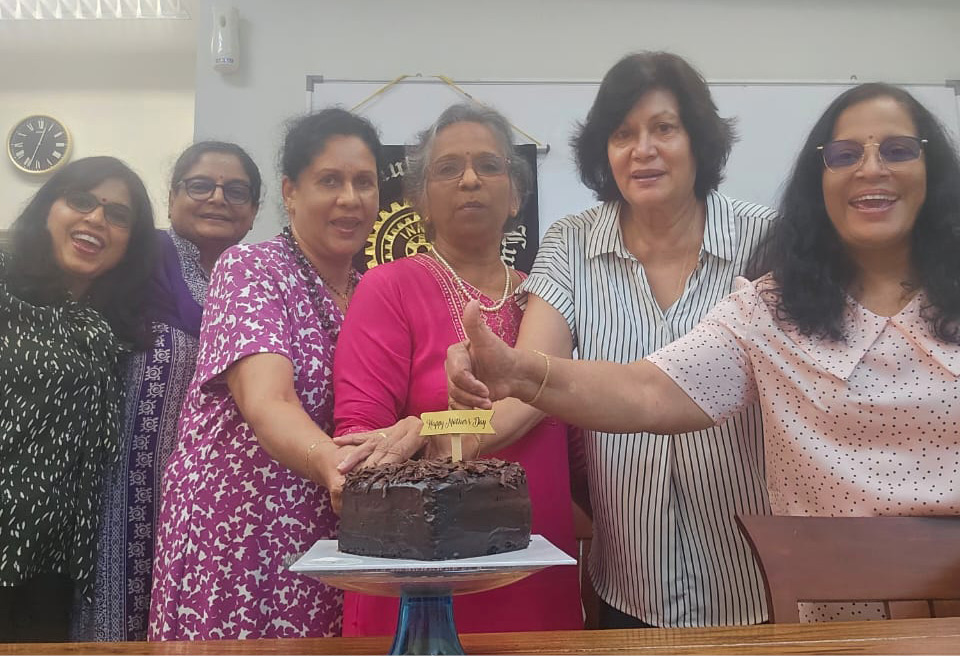 A group of six adults standing closely together indoors smiling and holding a chocolate cake with a candle on a table in front of them.
Description generated by AI