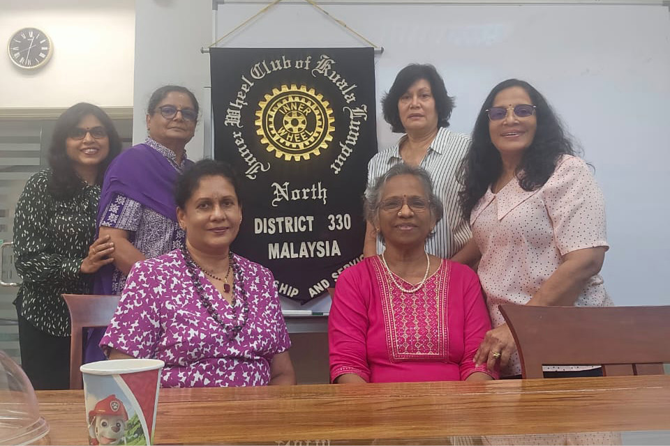 Six adults are gathered indoors around a table with a reflective surface, with a banner behind them displaying text related to a district in Malaysia.
Description generated by AI