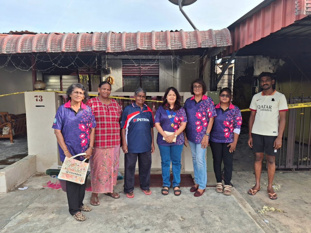 Seven adults stand together outdoors in front of a building, with five wearing matching purple shirts with pink floral patterns, one in a red plaid shirt and skirt, and one in a white sports shirt and shorts.

Description generated by AI