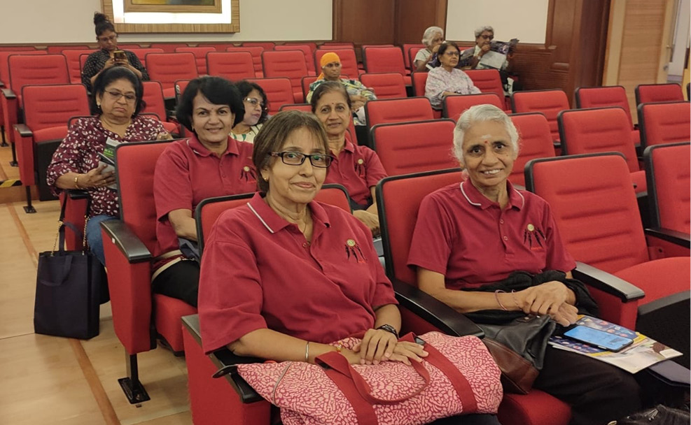 A group of adults wearing matching red shirts are seated in a conference room with red chairs, some holding bags and papers.
Description generated by AI