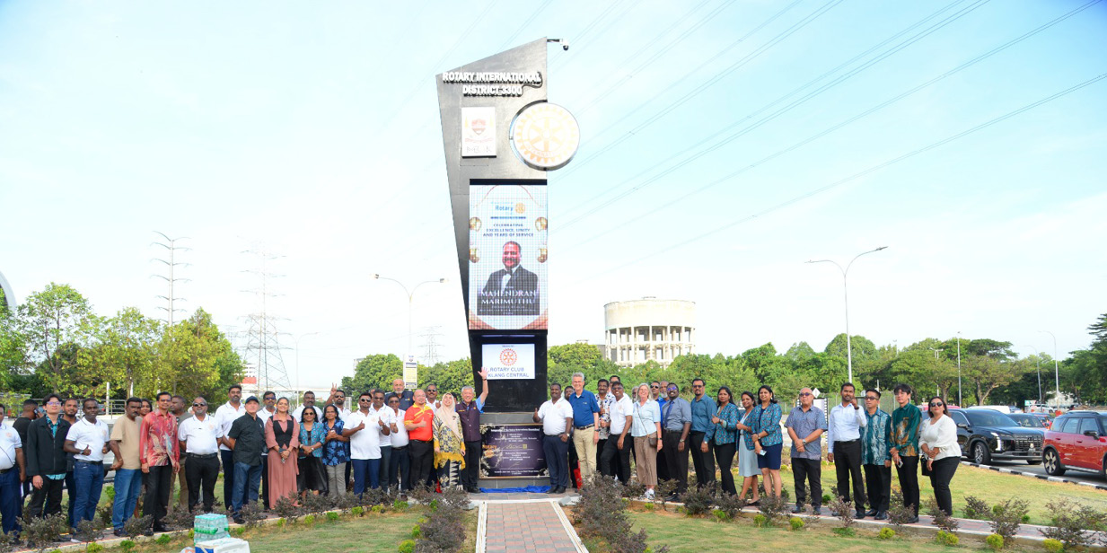 A large group of people stands outdoors on either side of a paved walkway in front of a tall signpost with banners and a clock, surrounded by grass and trees under a clear sky.

Description generated by AI