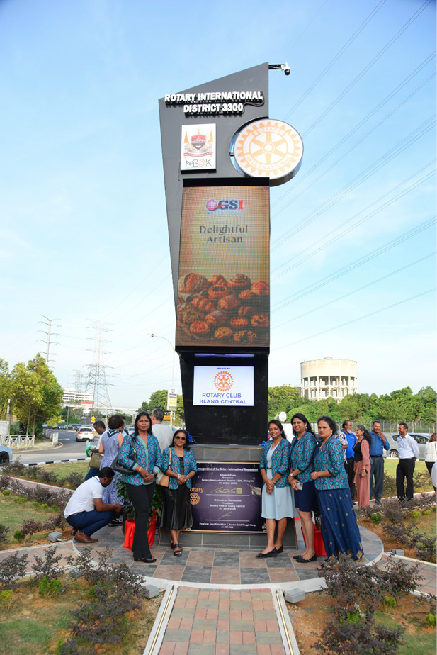 A group of people stands and kneels around a tall Rotary International District 3300 sign on a paved area near a road with trees and power lines in the background.

Description generated by AI