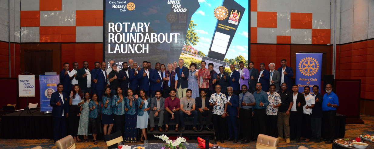 A large group of people dressed in formal and semi-formal attire stand on a stage in front of a screen displaying "Rotary Roundabout Launch" with tables set for a banquet in the foreground.

Description generated by AI