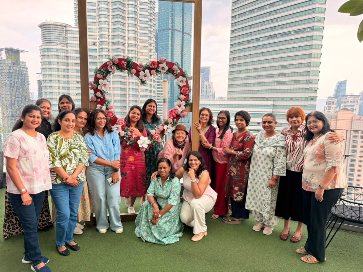A group of adults dressed in colorful and casual clothing pose together on a rooftop with a city skyline in the background and a large heart-shaped floral arrangement behind them.

Description generated by AI