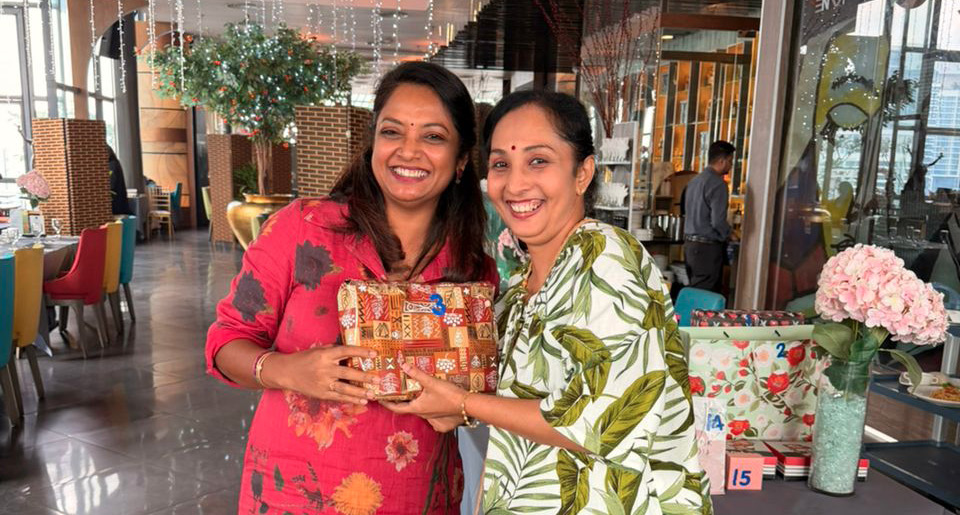 Two adults smiling and standing close together indoors, one wearing a red floral dress and white shoes holding a wrapped gift, and the other wearing a green and white patterned top with blue jeans.

Description generated by AI