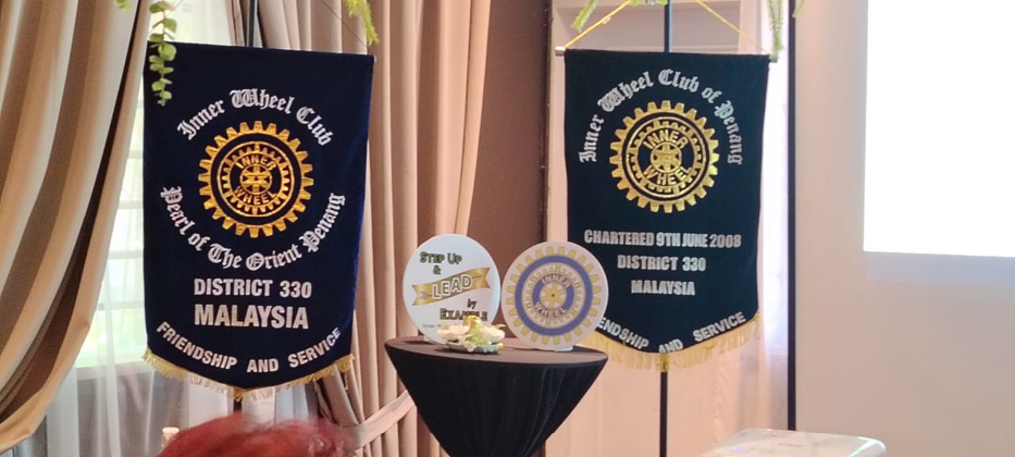 Two banners with the Rotary International emblem and text about the Inner Wheel Club of Penang are displayed hanging beside a small table with Rotary-themed decorations.
Description generated by AI
