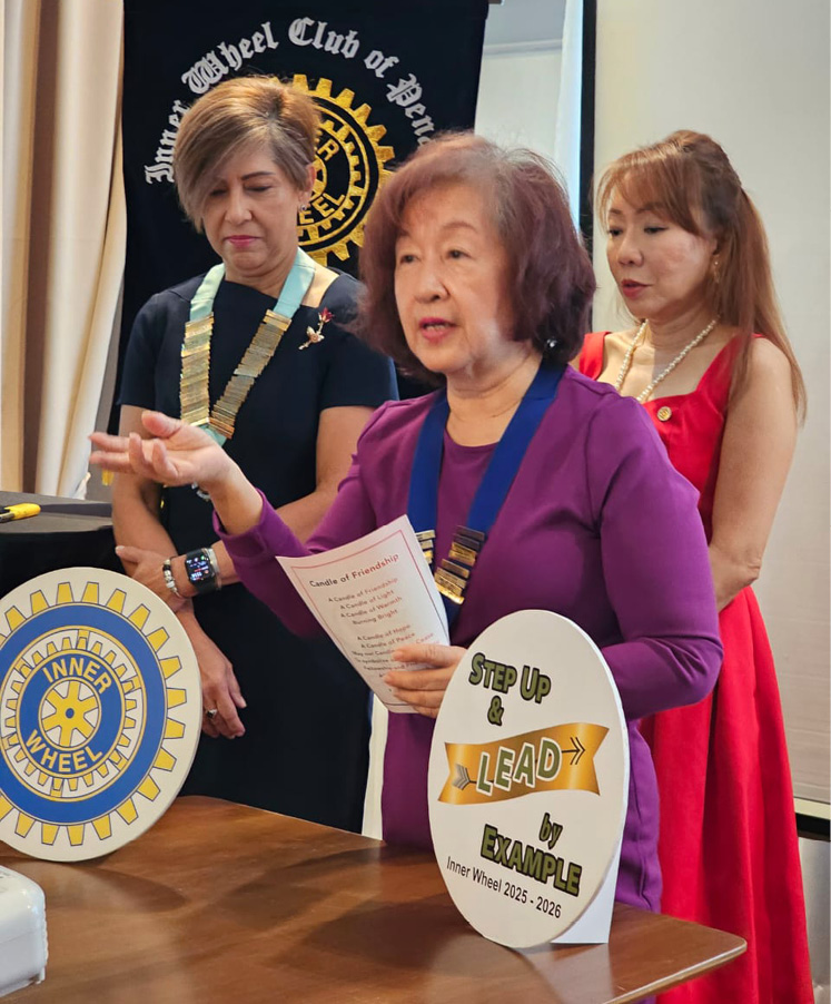 Three adults wearing ceremonial chains stand near a table with signs related to the Inner Wheel Club and a motivational message about leadership and example.
Description generated by AI
