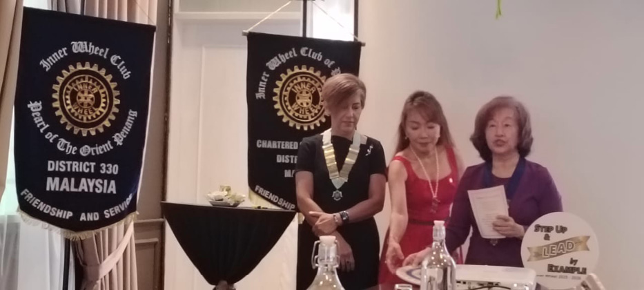 Three adults stand near a table with bottles and a sign, with two banners in the background indicating a Rotary Inner Wheel Club event in Malaysia.
Description generated by AI