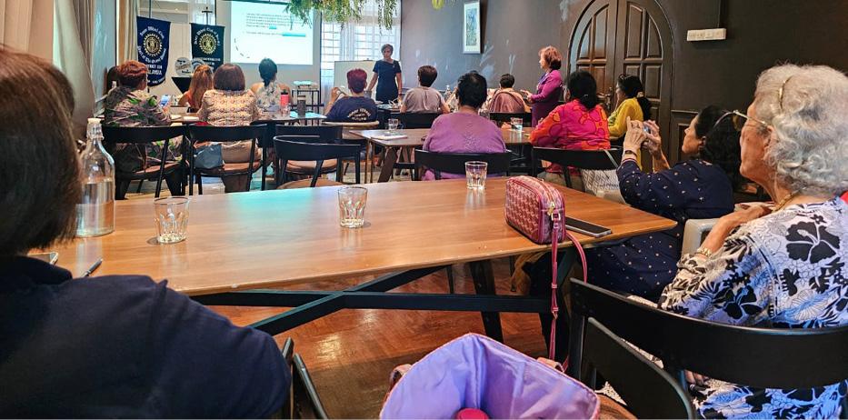 People seated at tables in a room attentively listening to a presenter speaking in front of a projection screen.
Description generated by AI