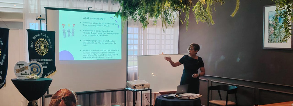 An adult wearing a black dress is giving a presentation to an audience in a dimly lit room with a projector screen displaying text and graphics.
Description generated by AI