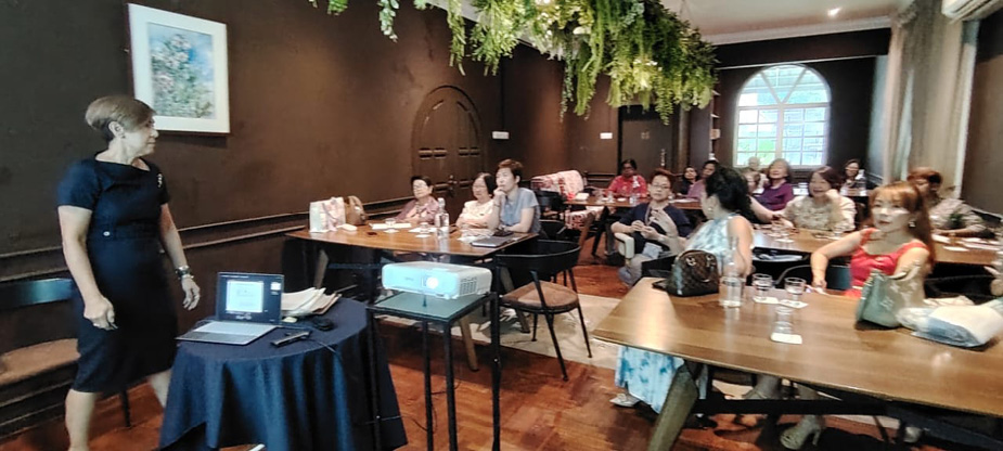A person in a dark dress is standing near a table with a laptop and projector while a group of people seated at tables attentively listen in a dimly lit room with hanging greenery.
Description generated by AI
