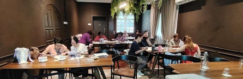 People seated around tables in a dimly lit room appear to be engaged in a meeting or workshop with a projector displaying content.
Description generated by AI