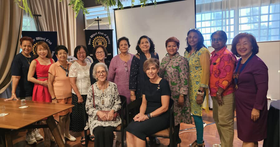 A group of people dressed in colorful and patterned clothing are standing and sitting together indoors in front of a screen and a banner, smiling for a group photo.
Description generated by AI