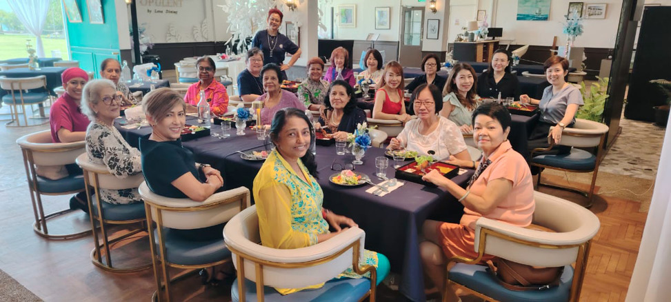 A group of adults sitting around a large table in a bright room are smiling and enjoying a meal together.
Description generated by AI