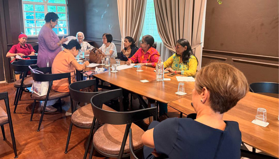 A group of people seated around a wooden table in a cozy room with hanging greenery and large windows, engaged in conversation and note-taking.
Description generated by AI
