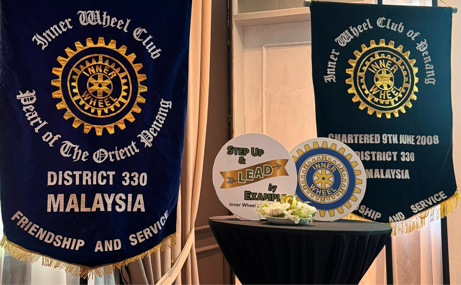Two dark blue banners with gold and white text and logos stand on either side of a small round table covered with a matching dark blue cloth tied with a gold ribbon, set against a backdrop of beige curtains and a white door.
Description generated by AI