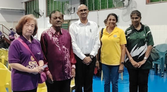 Five adults stand side by side indoors on a blue floor with yellow chairs and green windows in the background, wearing casual and semi-formal clothing.
Description generated by AI