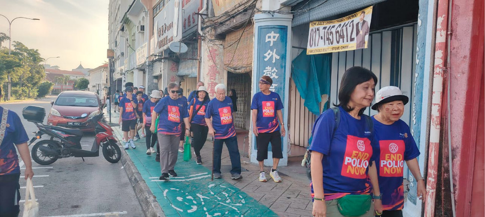 A group of people wearing matching blue shirts with "End Polio Now" printed on them are walking along a sidewalk in an urban area.
Description generated by AI