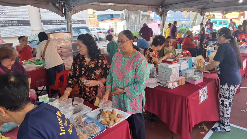 People in casual clothing are gathered under a tent at a market stall with tables covered in red cloth displaying various packaged food items and baked goods.
Description generated by AI