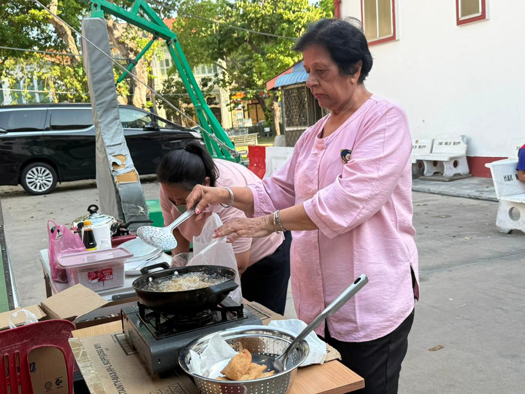 A person wearing a light pink shirt is cooking food in a pan on a portable stove outdoors while another person in a similar shirt is bent over nearby.
Description generated by AI