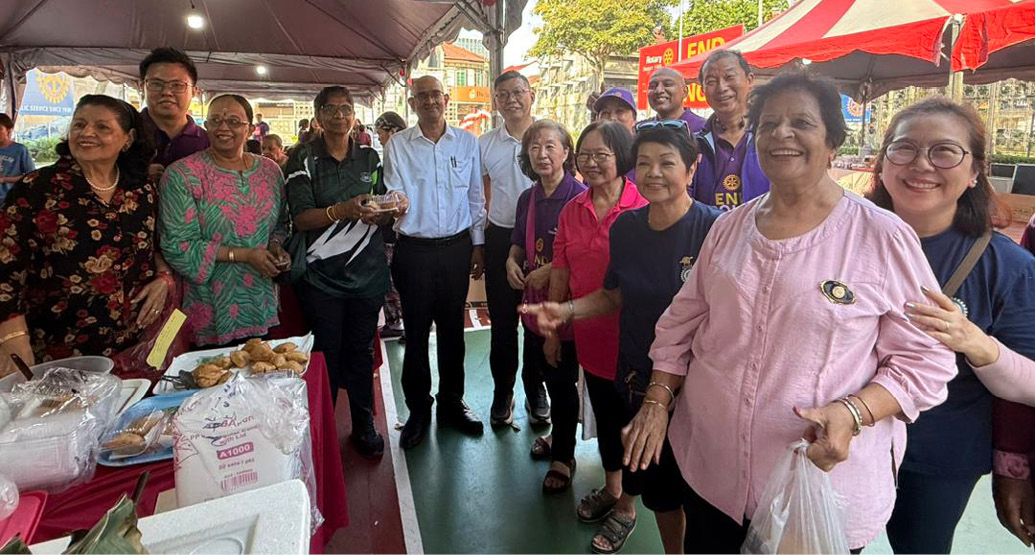 A group of people standing under a tent at an outdoor event with tables displaying food items and a red and white striped tent in the background.
Description generated by AI