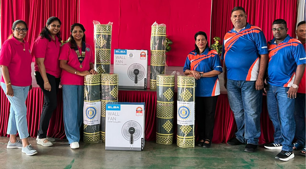 A group of people wearing matching pink and blue shirts stand smiling behind stacks of rolled mats and boxed wall fans in a room with red curtains.
Description generated by AI