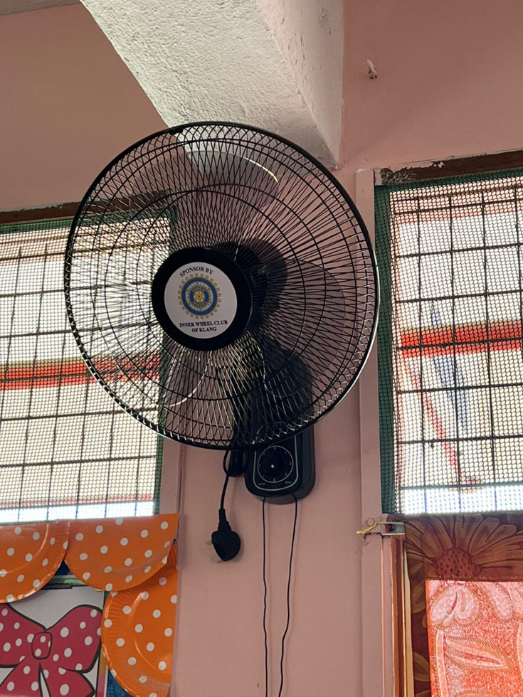 A black wall-mounted fan is attached to a pink wall between two windows with mesh screens and colorful curtains.
Description generated by AI