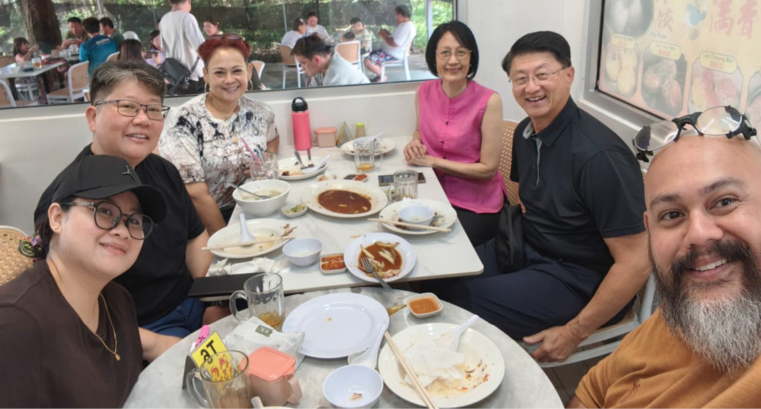 A group of seven people sit around a table with plates of food and drinks in a casual dining setting, smiling and engaging with each other.

Description generated by AI
