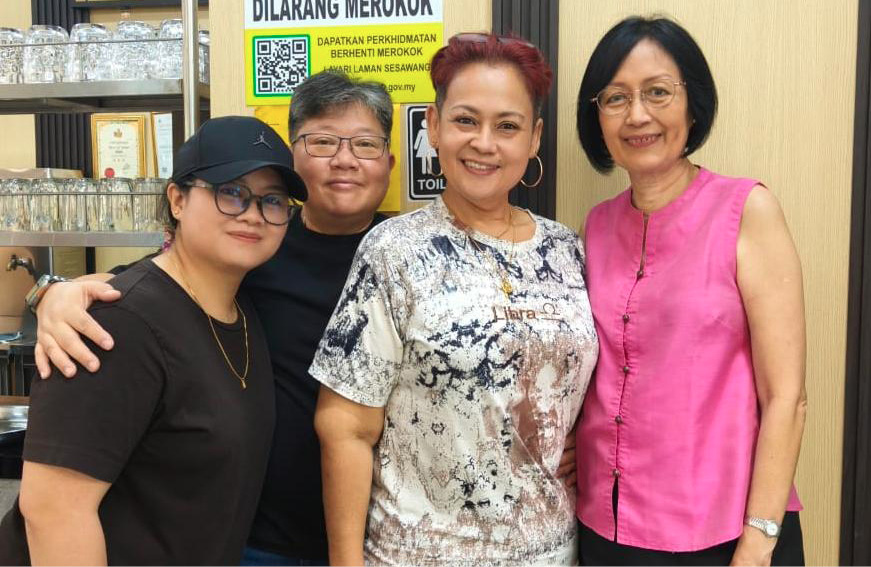 Four adults stand closely together indoors smiling, with one wearing a black cap and glasses, another in a pink sleeveless top, and a sign with text in the background.

Description generated by AI