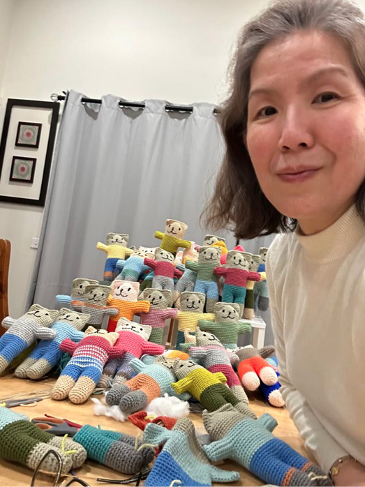 A person with shoulder-length hair wearing a light-colored sweater is sitting at a table filled with numerous colorful knitted dolls arranged in rows.

Description generated by AI