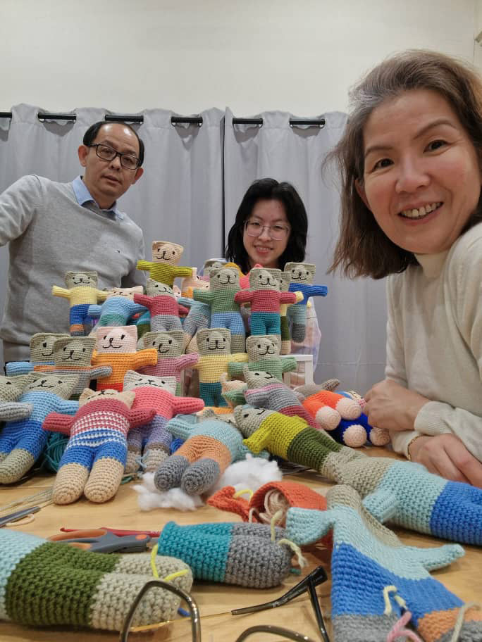 Three adults smile and pose behind a table filled with colorful crocheted dolls and yarn crafts in a cozy indoor setting.

Description generated by AI