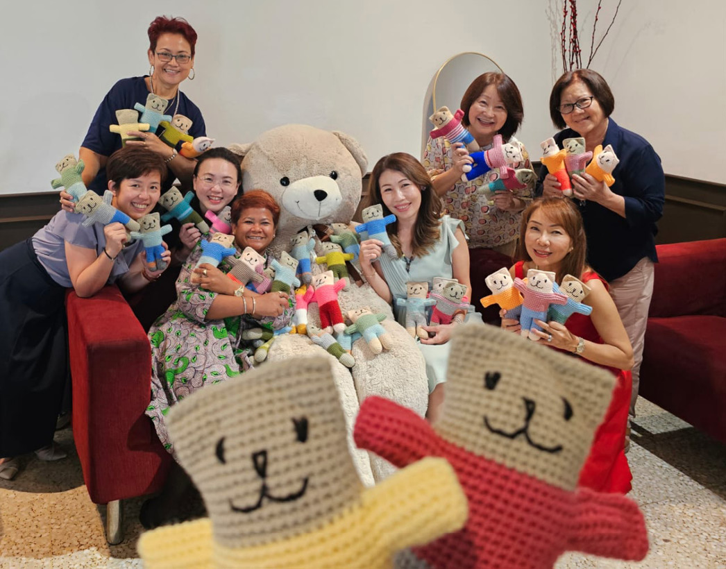 A group of eight adults smiling and holding colorful knitted bear dolls while sitting and standing around a large teddy bear in a cozy indoor setting.

Description generated by AI