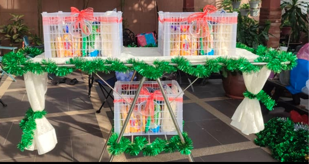 Three gift baskets wrapped with red ribbons are displayed on a table decorated with green garlands and white curtains tied with green garlands in an outdoor setting.
Description generated by AI