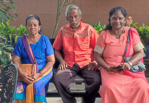 Three seniors wearing colorful traditional clothing are sitting on a bench outdoors with plants and a building in the background.
Description generated by AI