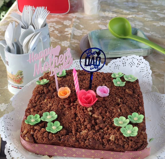 A heart-shaped chocolate cake decorated with small green flowers and pink roses has candles and a "Happy Mother's Day" topper on a table with a floral tablecloth, alongside a Coca-Cola cooler, plastic cups, and utensils.
Description generated by AI